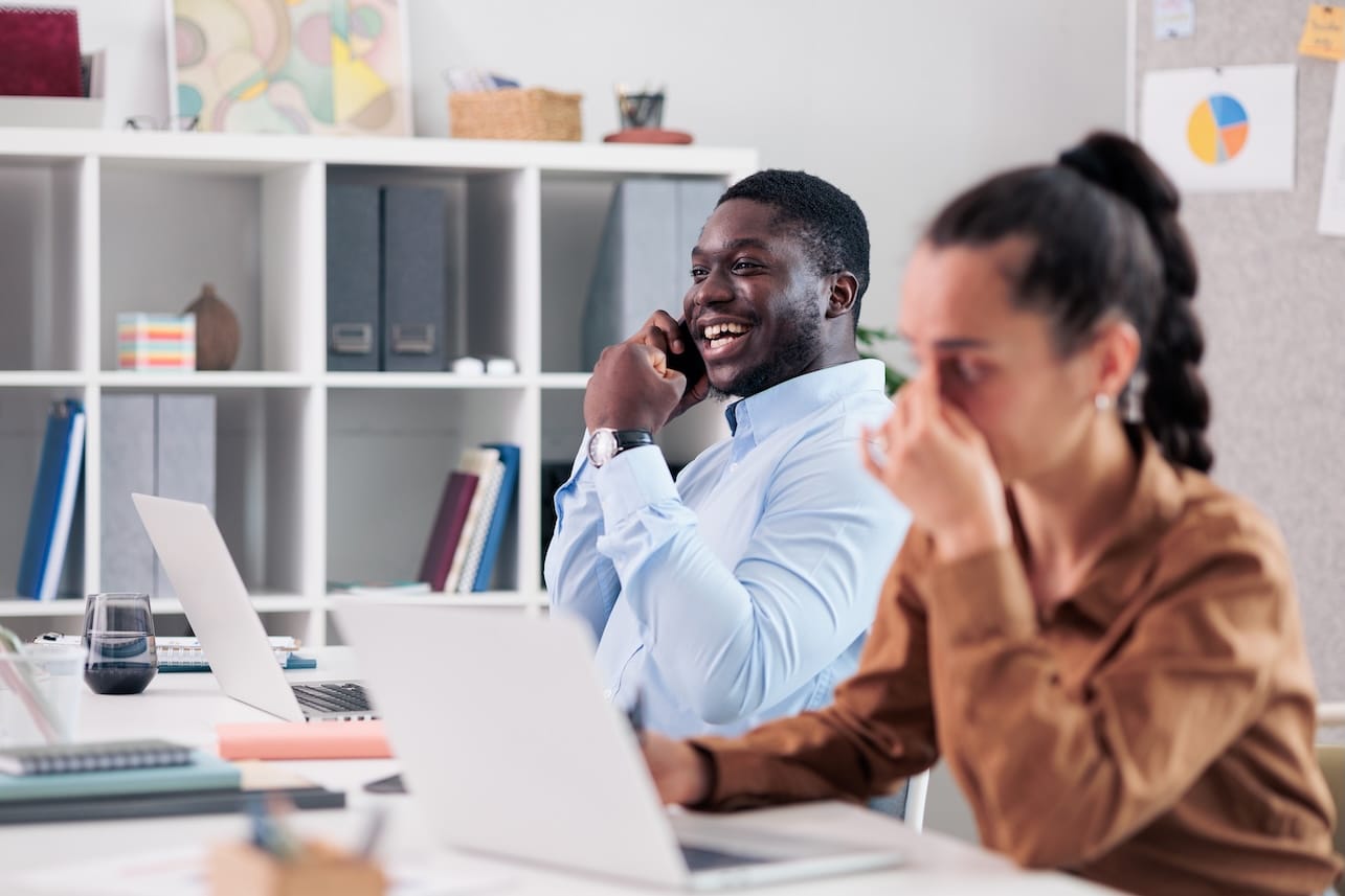 Man on a phone call laughing loudly while woman coworker tries to focus in an open-plan office