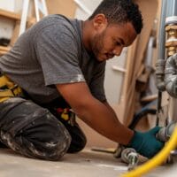 A male worker kneels, wearing a grey shirt, yellow tool belt, and dark work pants, while working on plumbing fixtures and pipes in a construction or repair setting. The scene shows a close-up view of his hands and the work.